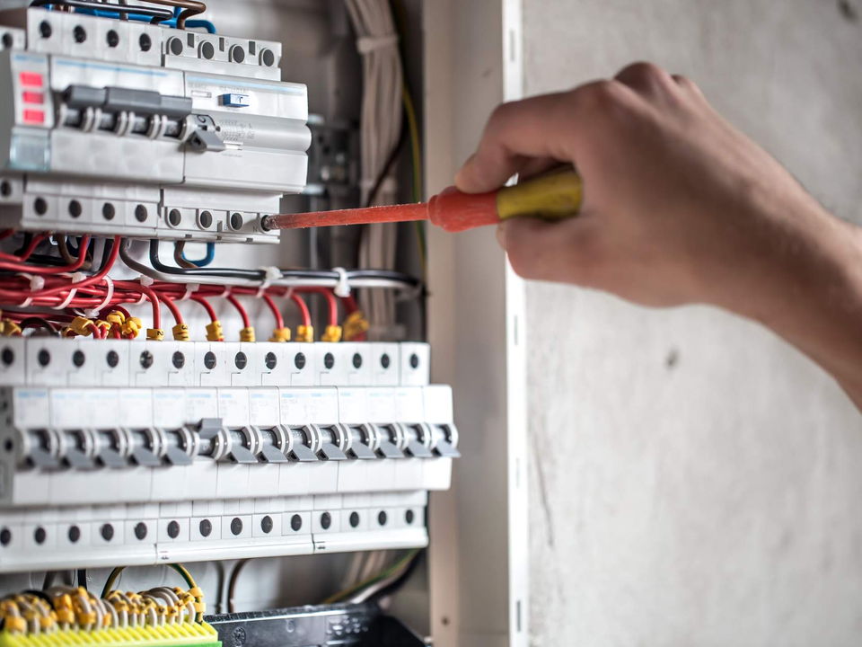 A hand with a red-handled screwdriver tightening a terminal in a panel of circuit breakers