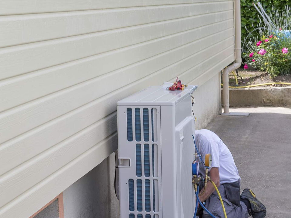 A technician kneeling beside a heat pump outdoor unit against the weatherboard wall of a house