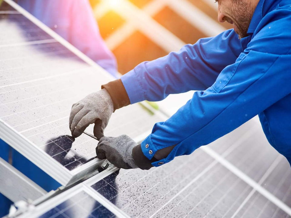 An electrician in blue overalls and gloves fastening a solar panel to its rail in bright sunlight