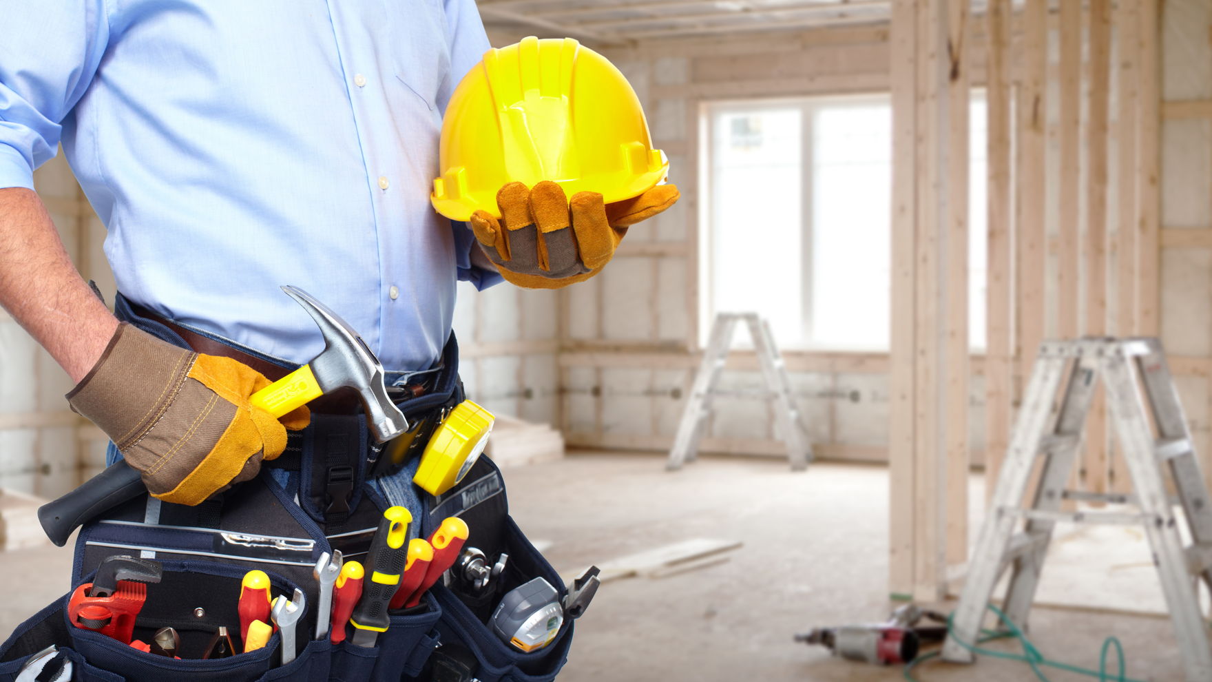 A tradesman with a tool belt of screwdrivers and pliers holding a yellow hard hat, in a timber-framed house under construction