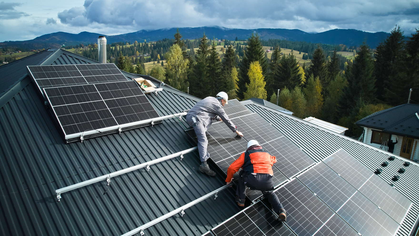 Two installers fixing solar panels to mounting rails on a long-run metal roof, with forest and hills behind