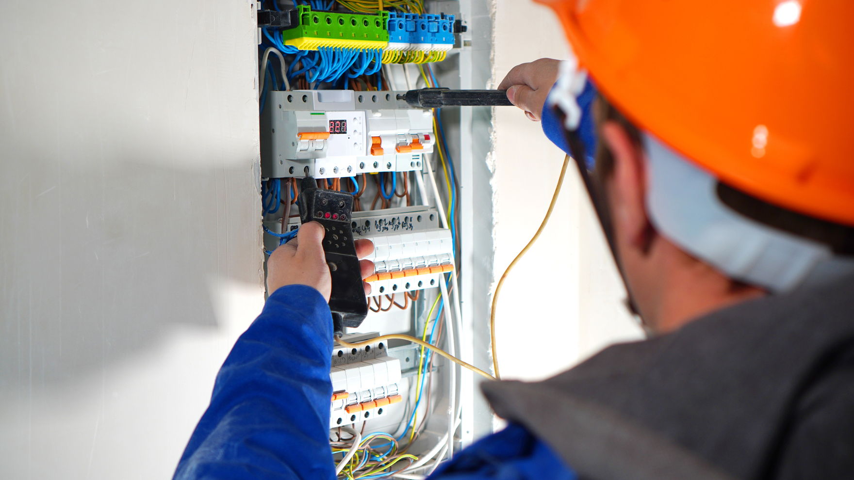 An electrician in a blue shirt and orange hard hat testing circuit breakers in a wall-mounted switchboard